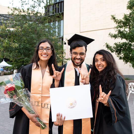 Three graduates pose with diploma and give hook em horns